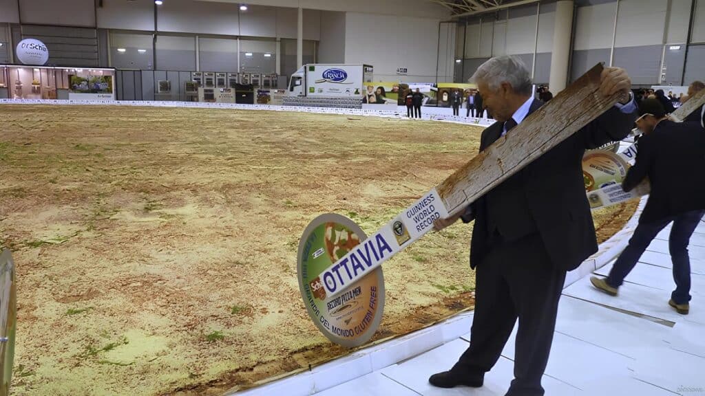 World's largest pizza in exhibition with people holding giant pizza cutter sign labeled Ottavia for Guinness World Record.