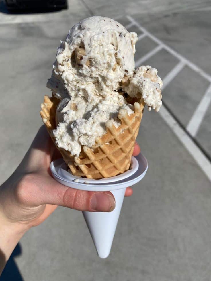 Hand holding a scoop of creamy ice cream in a waffle cone with cone holder, outdoors on a sunny day.