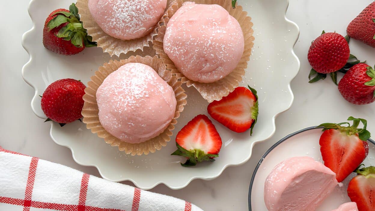 Pink strawberry mochi on a plate with fresh strawberries and a red-checkered napkin on the side.