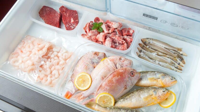 Variety of frozen seafood and meats in a freezer, including fish, shrimp, steaks, and sliced meat with lemon slices.