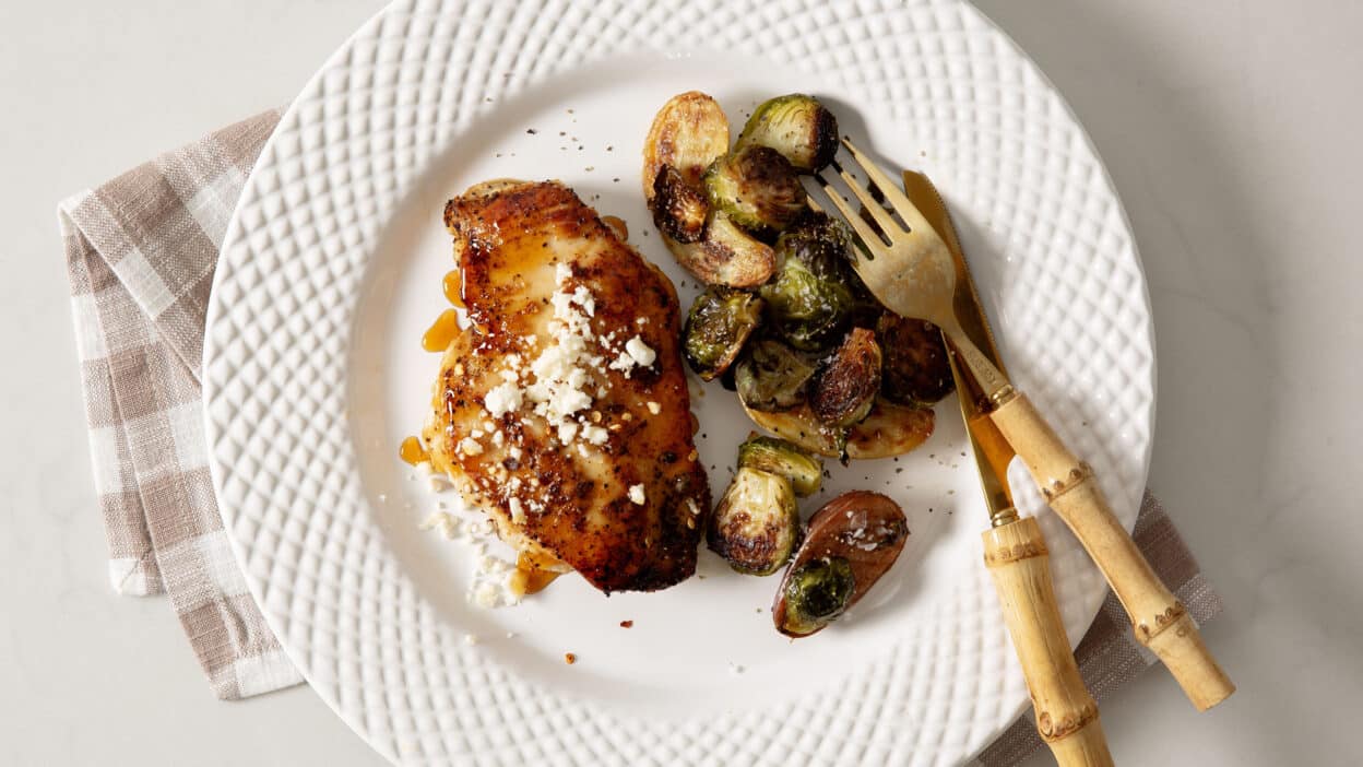 Grilled chicken breast with feta and roasted Brussels sprouts on a white plate with a bamboo fork and knife.