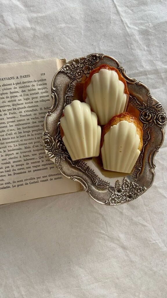 Elegant silver platter with madeleines next to an open book on linen fabric.