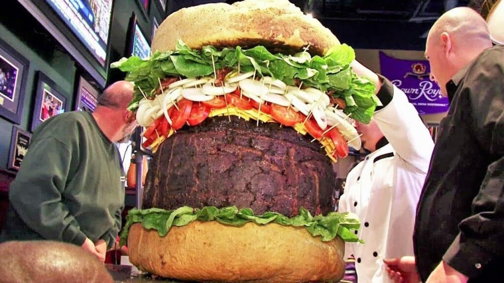 Giant burger challenge with layers of lettuce, onions, tomatoes, and massive beef patty in a lively restaurant setting.