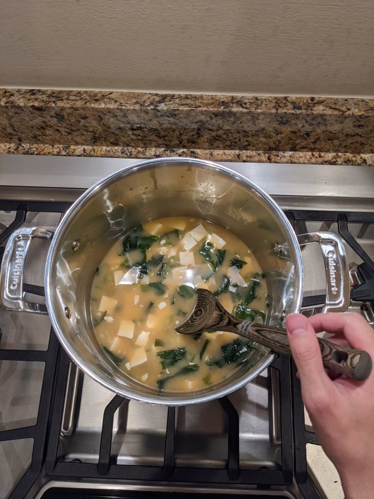 Hand stirring miso soup with tofu and greens in a pot on a stove. Home-cooked comfort food.