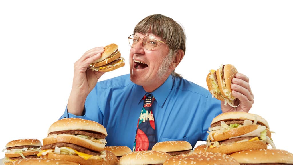 Man in blue shirt happily holding and surrounded by burgers, wearing a colorful tie.