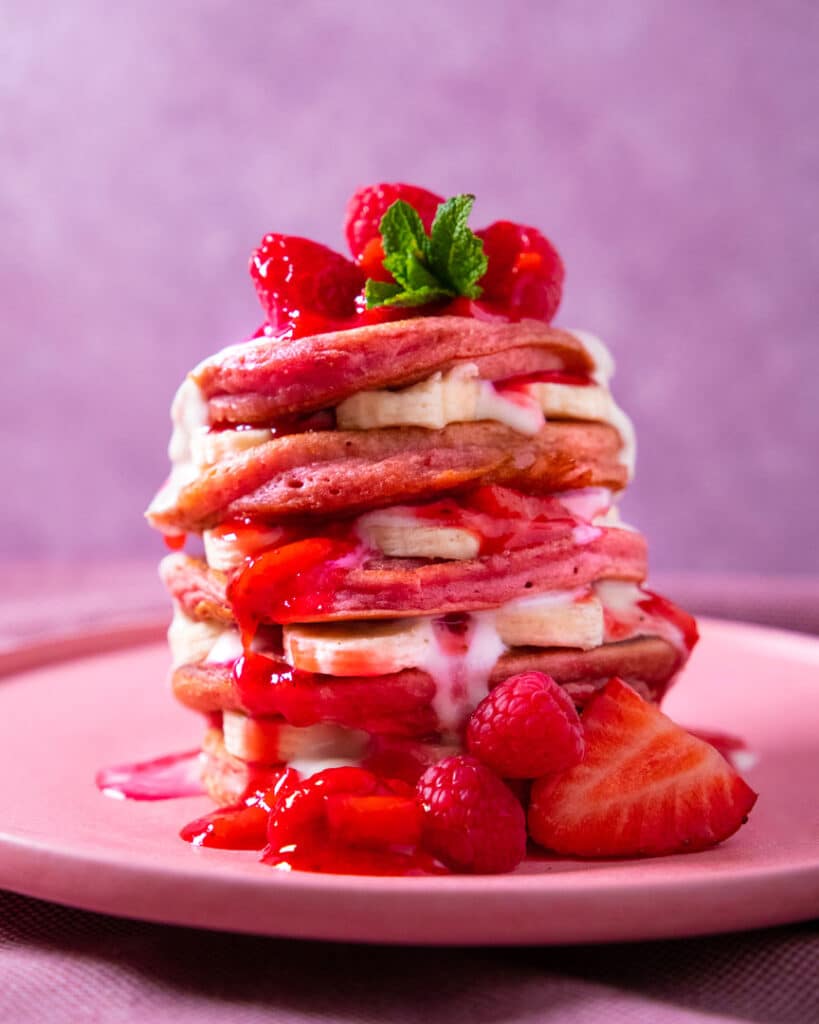 Stack of pink pancakes with bananas, raspberries, strawberry sauce, and mint on a pink plate. Perfect for breakfast delight.