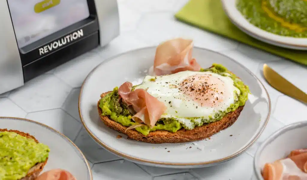 Avocado toast with poached egg and ham on a plate next to a toaster in a bright kitchen setting.