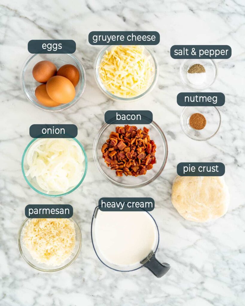 Labeled quiche ingredients on a marble surface: eggs, cheeses, bacon, onion, heavy cream, pie crust, spices.