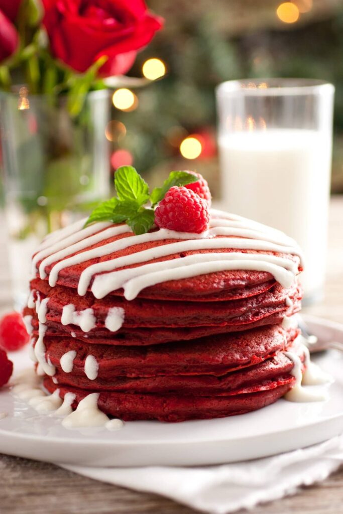 Stack of red velvet pancakes with cream drizzle, topped with raspberry and mint, served with milk.