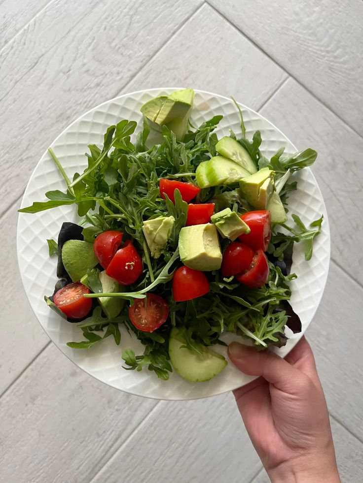 Fresh salad with cherry tomatoes, avocado, cucumber, and arugula on a white plate, held by a hand on tile background.