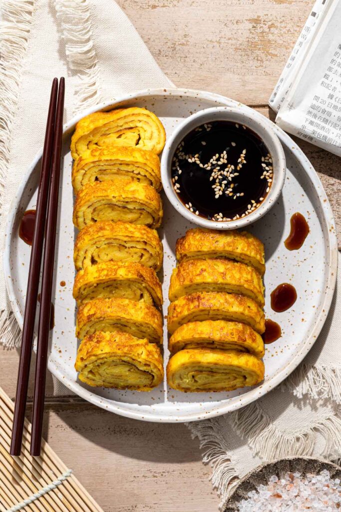 Japanese rolled omelette slices with soy dipping sauce, garnished with sesame seeds, served on a white plate.