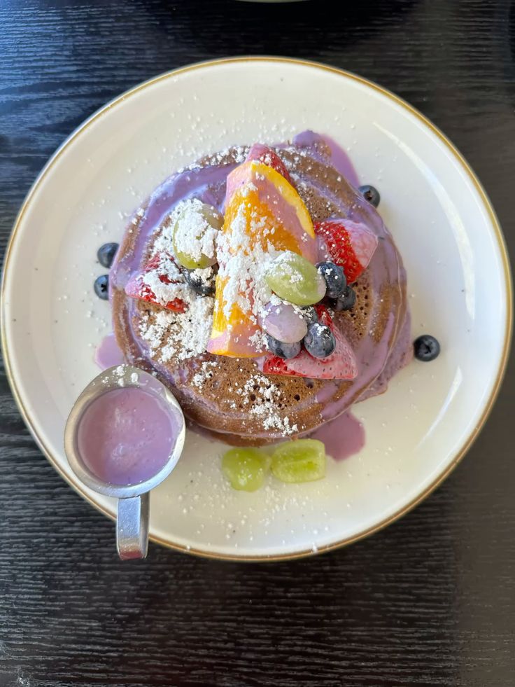 Vibrant pancakes with fruit toppings and purple sauce, garnished with powdered sugar, served on a white plate.