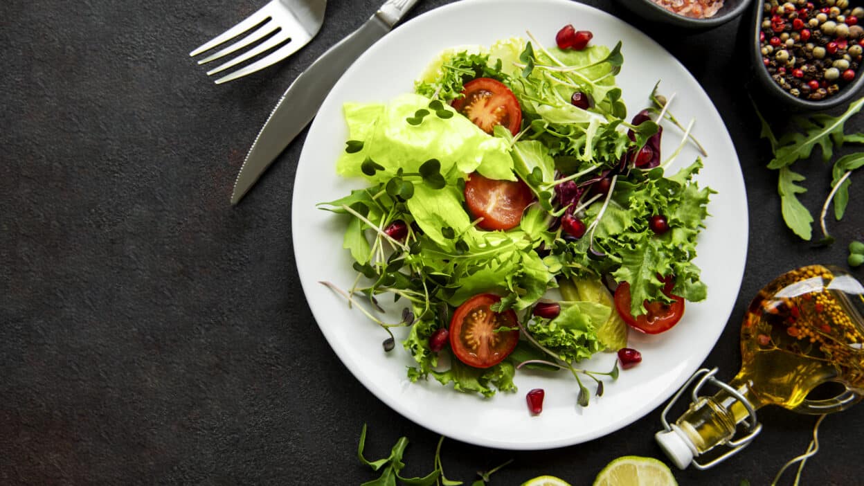 Fresh salad with lettuce, cherry tomatoes, and pomegranate seeds on a white plate, surrounded by seasonings and lime.