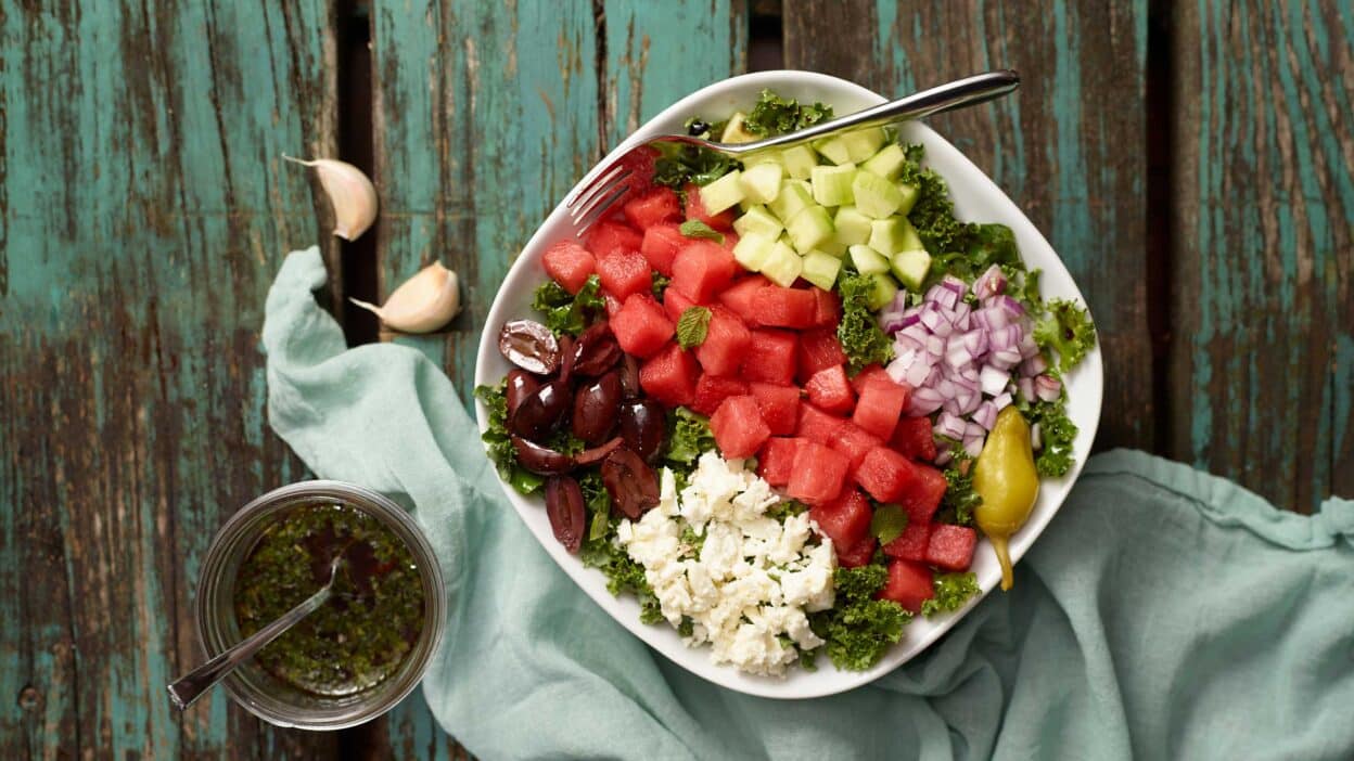 Fresh Greek salad with watermelon, cucumber, feta, onion, olives, and dressing on a rustic wooden background.