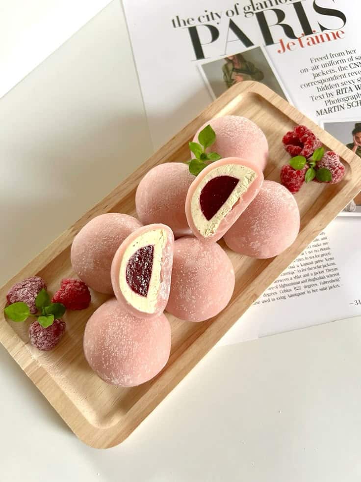 Pink mochi filled with raspberry cream on a wooden tray, garnished with fresh raspberries and mint leaves.
