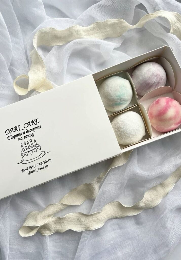 Box of multicolored mochi desserts in packaging on a white fabric background, featuring ribbon detail.