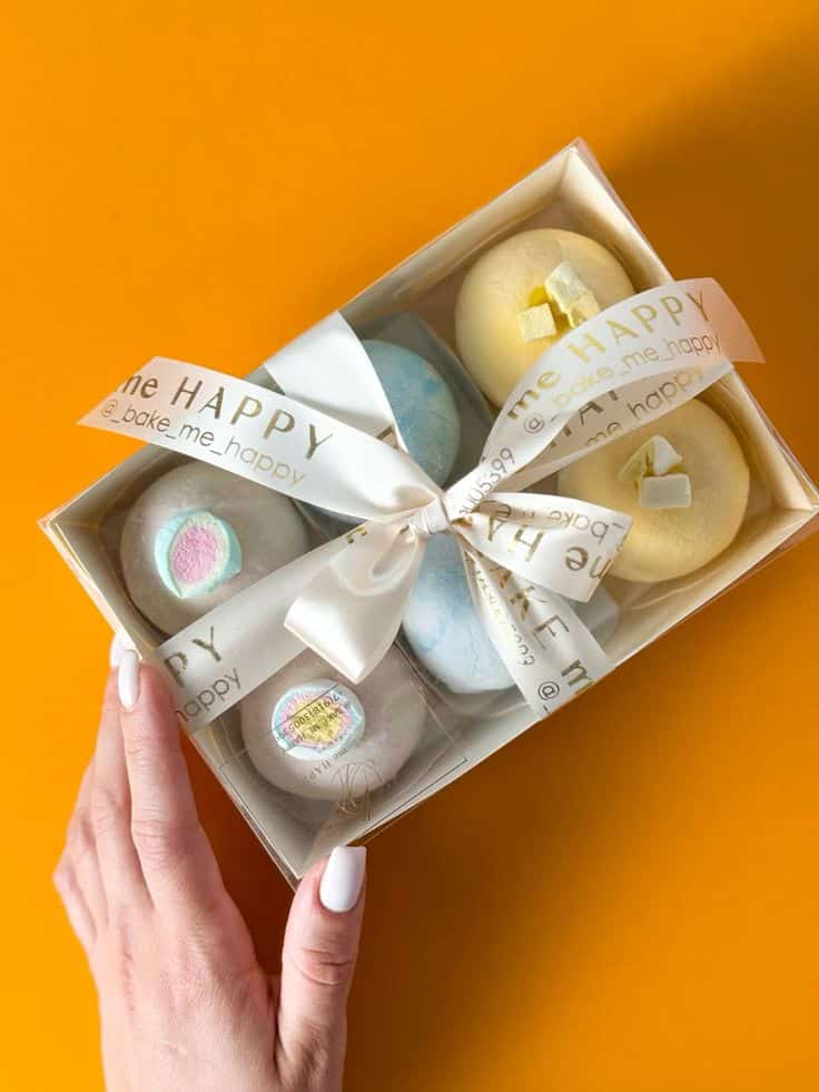 Box of colorful bath bombs with decorative ribbon, held by a hand on an orange background.