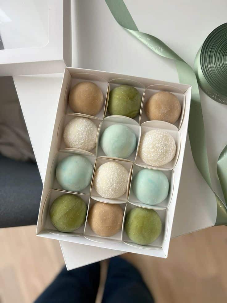 Assorted pastel mochi set in a white box, tied with a green ribbon, on a table.