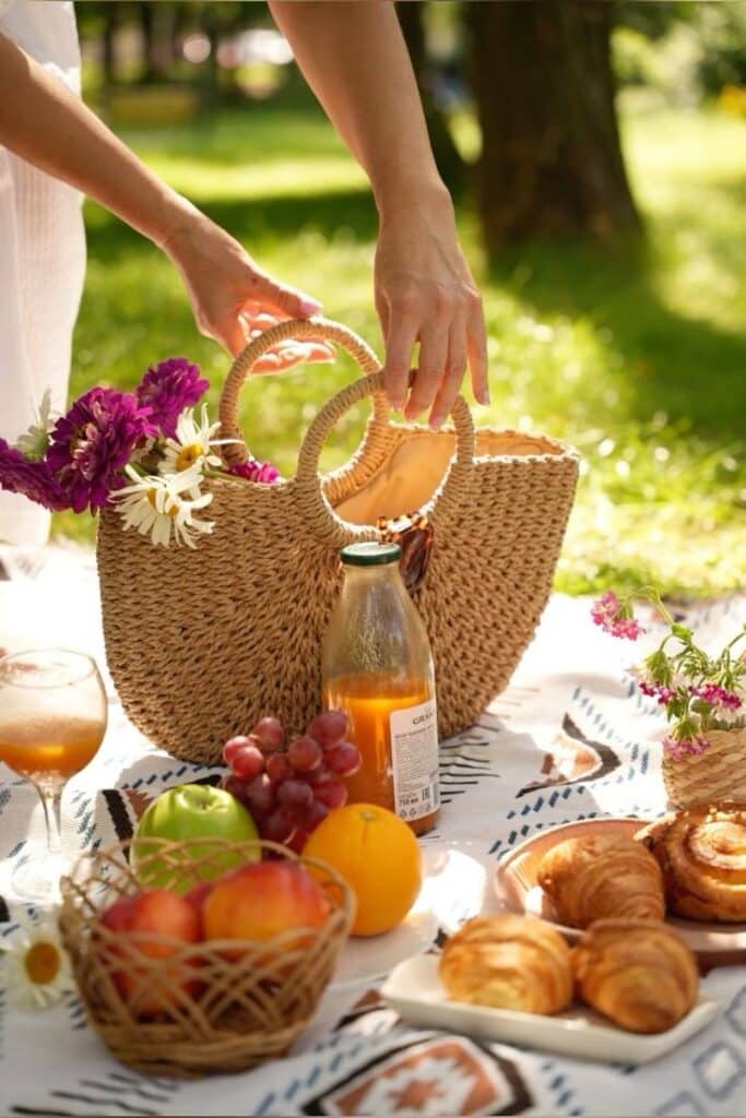 Summer picnic with fruit, flowers, and juice on a patterned blanket. A woven basket adds a rustic touch.