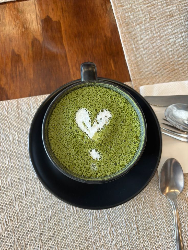 Matcha latte with heart design on wooden table, black cup, and saucer, alongside spoon and napkin.