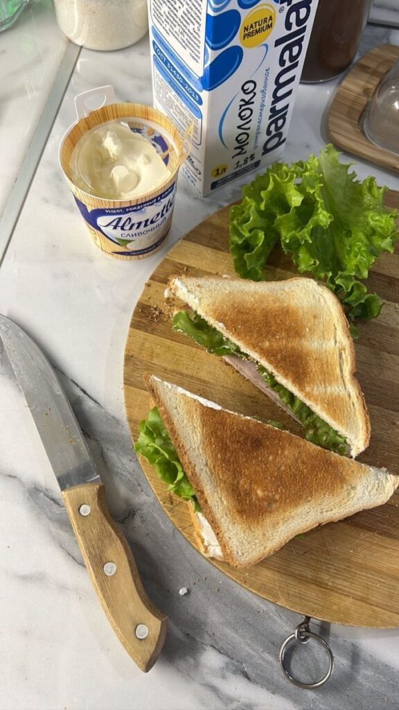 Fresh lettuce sandwich on a wooden board with cream cheese, knife, and milk carton on marble counter.