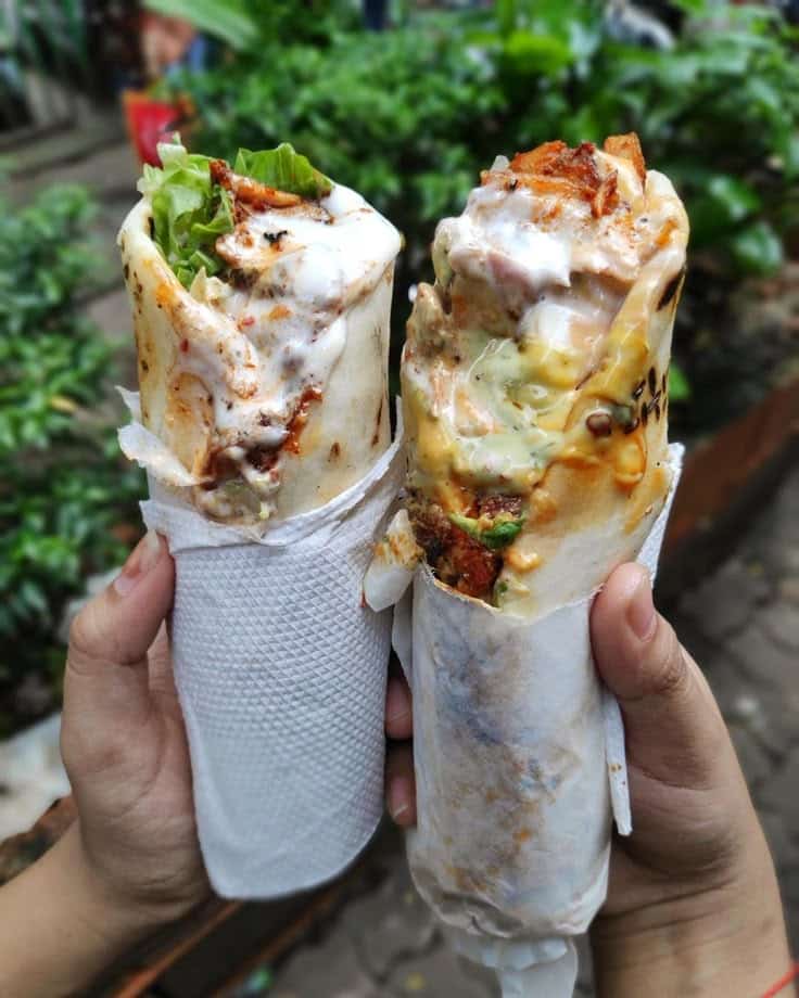 Two delicious stuffed wraps with sauces held in hands, wrapped in white paper towels, outdoors.