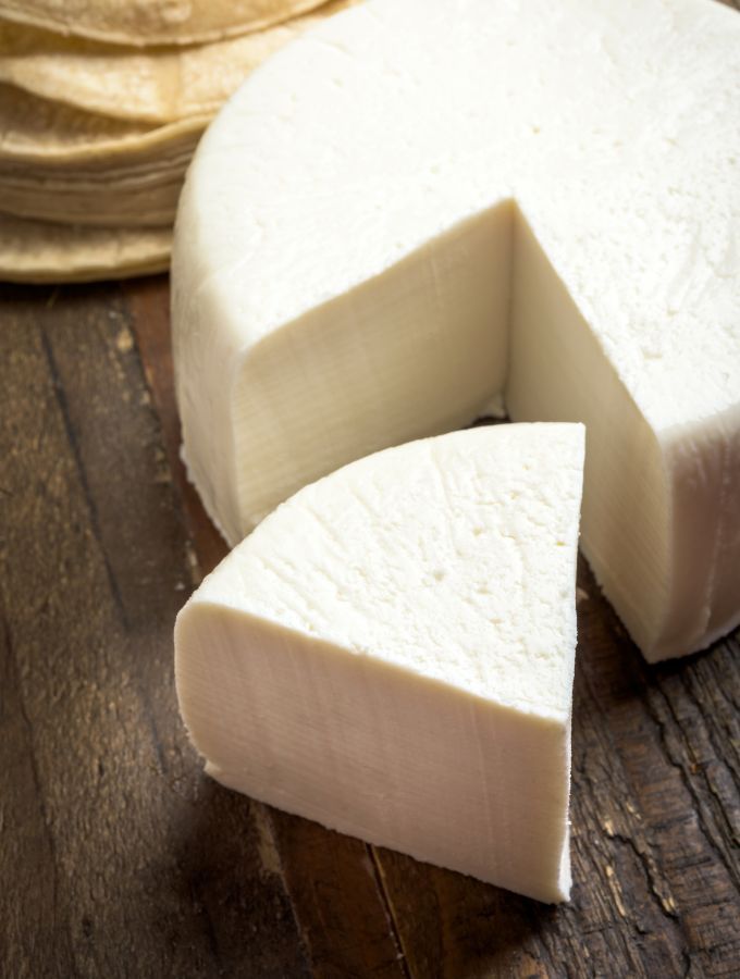 Round queso fresco cheese wheel with a slice cut out, placed on a rustic wooden table.