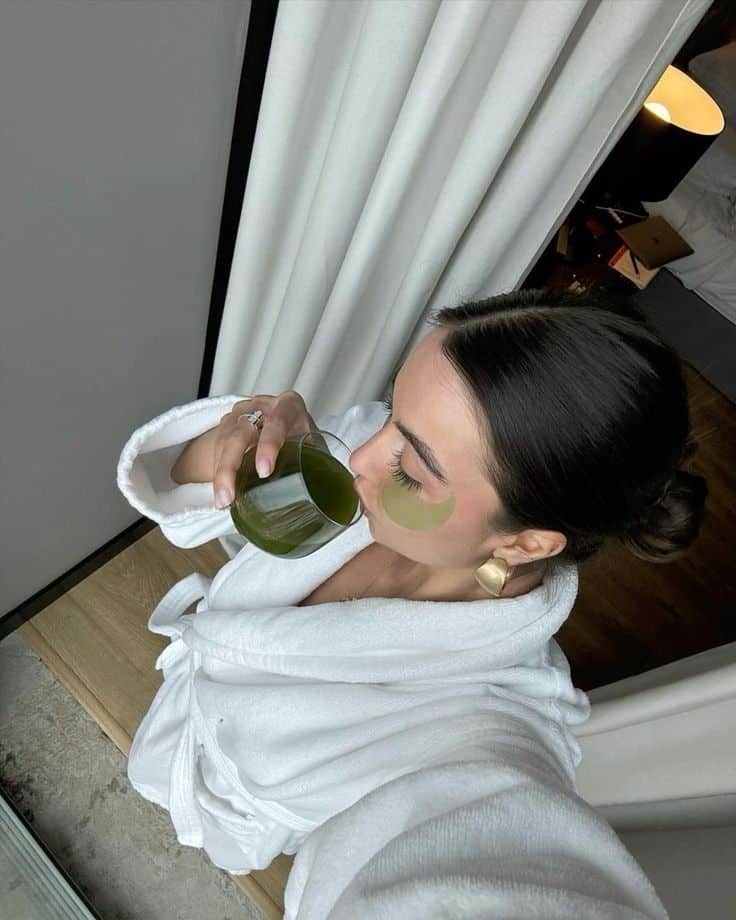 Woman in a white robe enjoying green drink with under-eye patches for skincare relaxation.