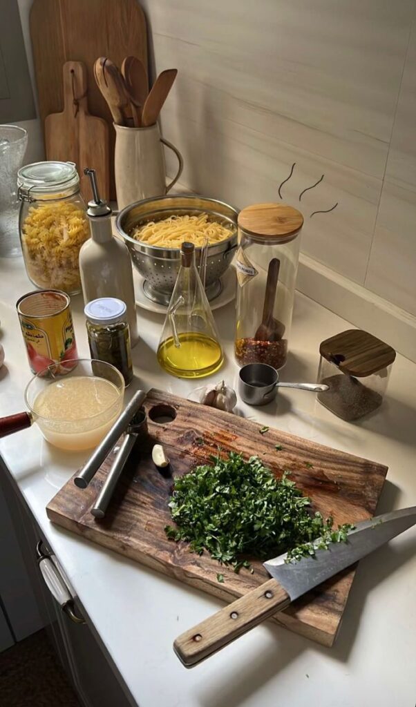 Kitchen counter with pasta ingredients: chopped herbs, garlic, olive oil, pasta, and cutting board for cooking prep.