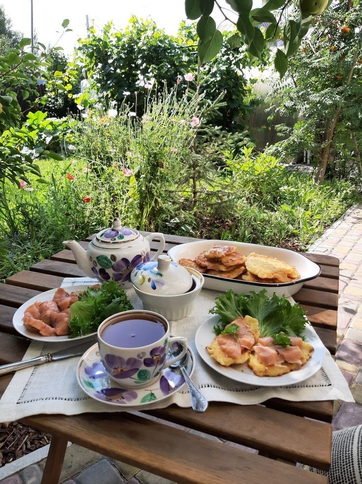 Garden tea party with floral china set, pancakes, salmon, and greenery on a wooden table surrounded by lush plants.