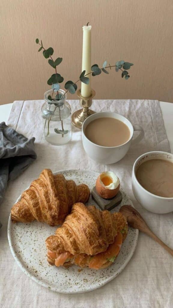 Delicious breakfast with croissants, soft-boiled egg, coffee, vase of greenery, and candle on a table setting.
