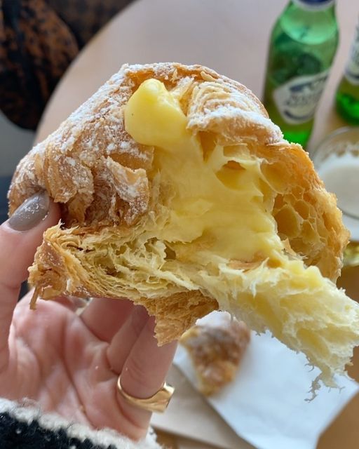 Close-up of a hand holding a flaky croissant filled with creamy custard, with bottles in the blurry background.