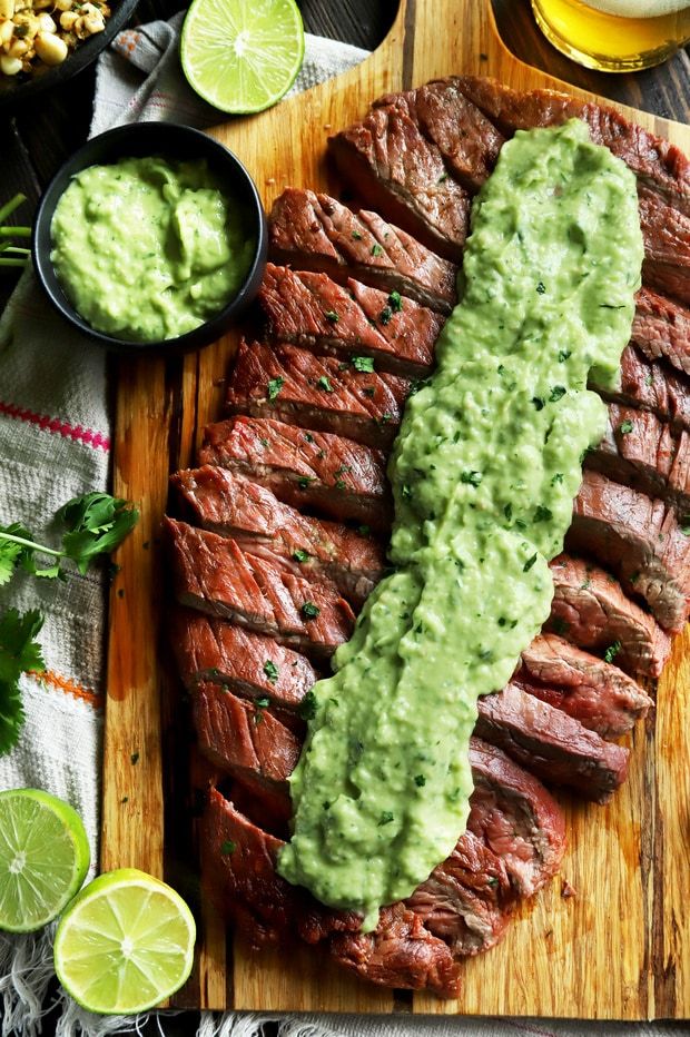 Grilled steak topped with creamy avocado sauce on a wooden board, garnished with lime slices.