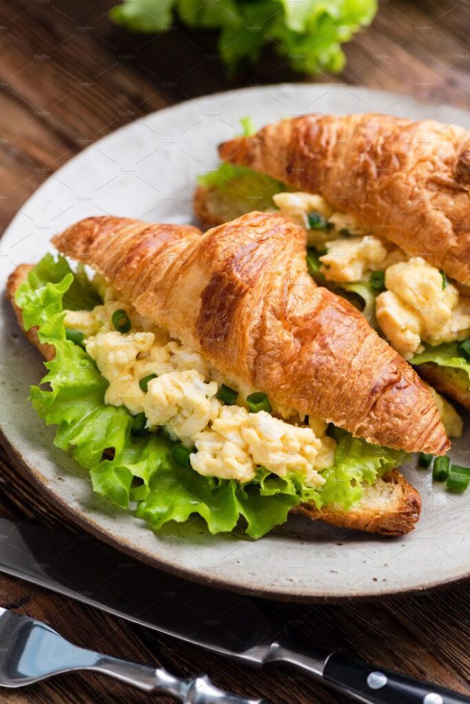 Croissant sandwich with scrambled eggs, fresh lettuce, and green onions on a plate, ideal breakfast choice.