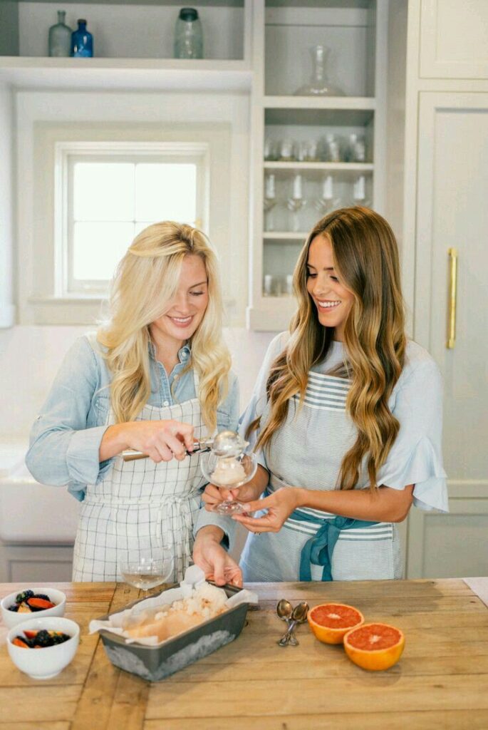 Two women making homemade ice cream in a bright kitchen, surrounded by fruit and bowls.