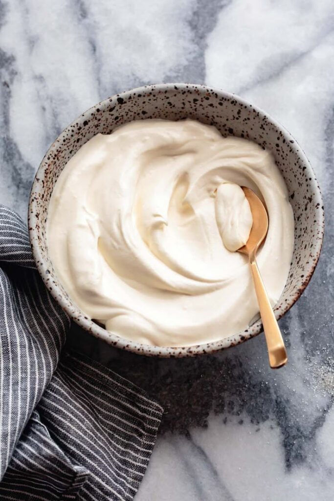 Creamy homemade yogurt in a speckled bowl with a golden spoon on a marble surface, striped cloth beside.
