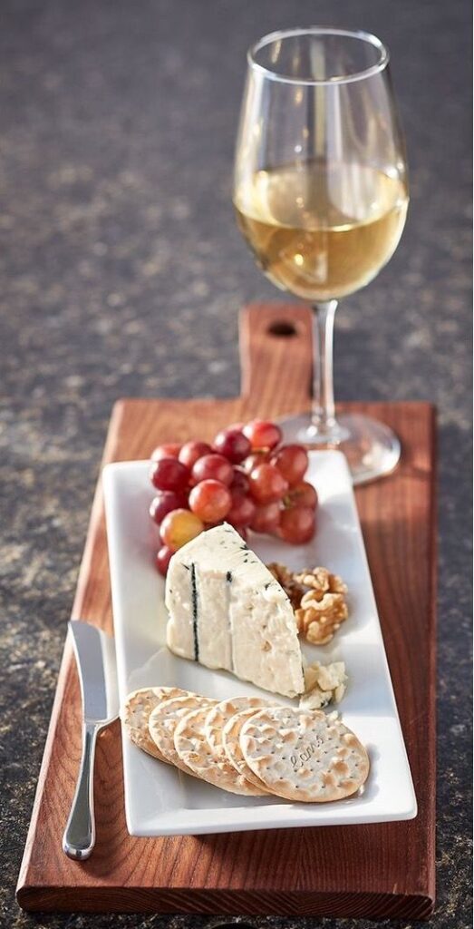 Cheese platter with crackers, grapes, walnuts, and a glass of white wine on a wooden board.