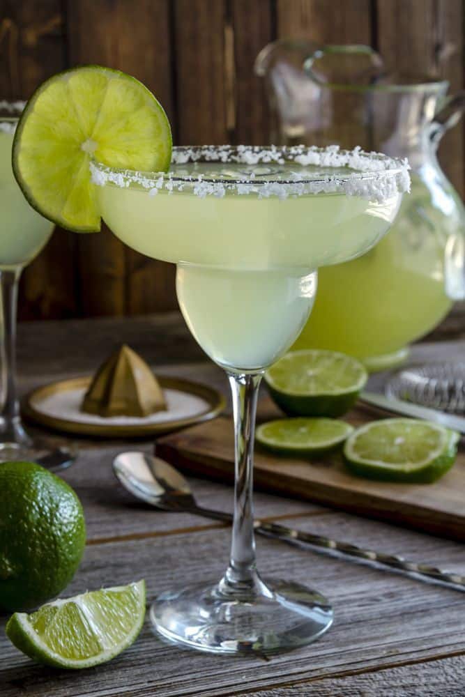 Refreshing margarita with lime garnish and salt rim on rustic table, surrounded by sliced limes and pitcher.