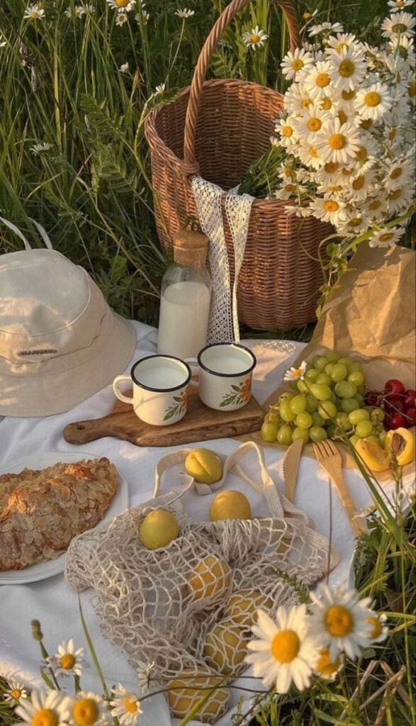 Summer picnic setup with daisies, wicker basket, fruits, bread, and mugs on a white cloth in a grassy field.
