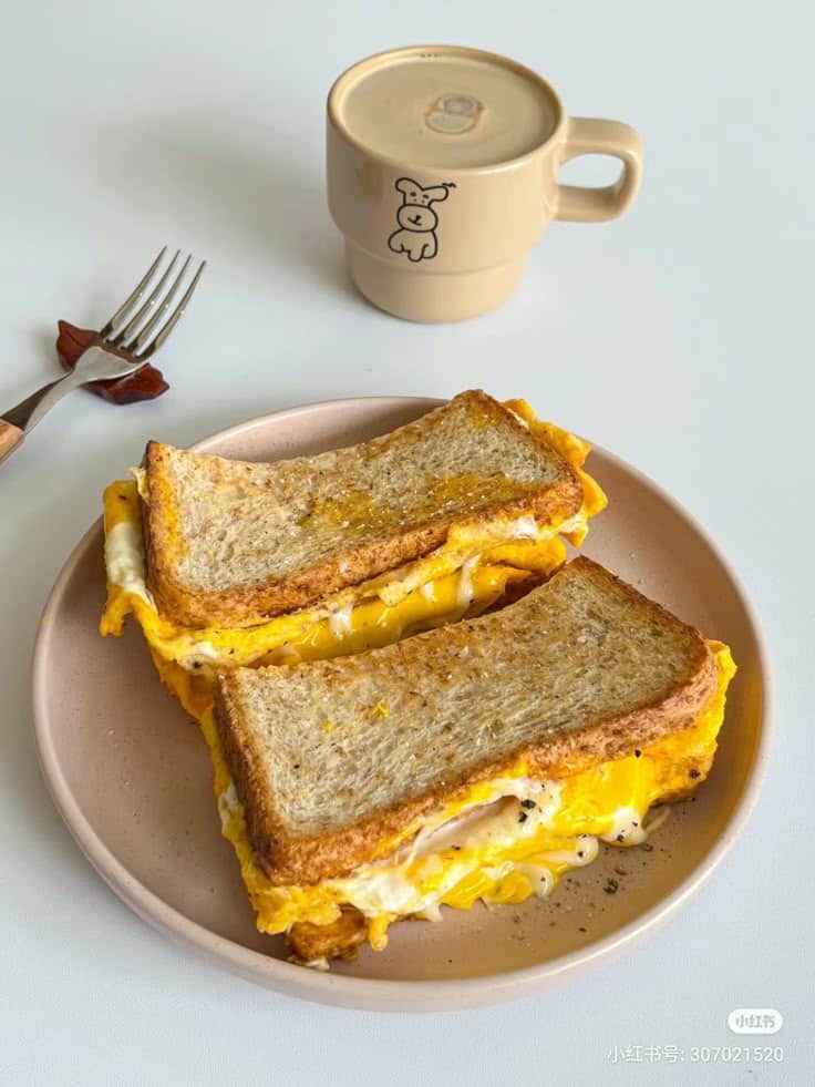 Grilled cheese sandwiches with eggs on a plate, served with a cute mug of coffee and fork on the side.