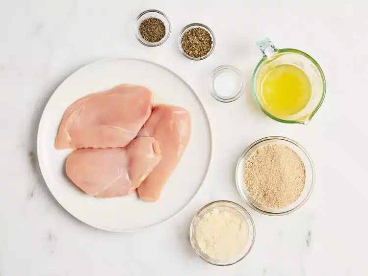 Raw chicken breasts with seasoning, breadcrumbs, cheese, and melted butter on a marble surface, ready for cooking.