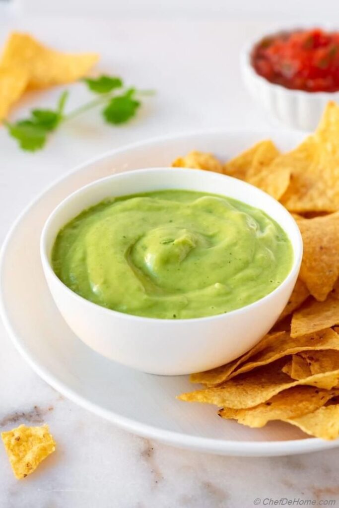 Bowl of creamy guacamole with tortilla chips on a white plate, garnished with fresh cilantro, perfect for parties.
