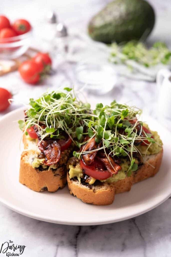 Open-faced avocado toast with bacon, tomato, and sprouts on a white plate, surrounded by fresh ingredients.