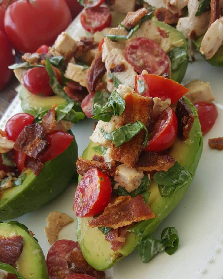 Avocado boats topped with cherry tomatoes, chicken, spinach, and crispy bacon on a white plate.