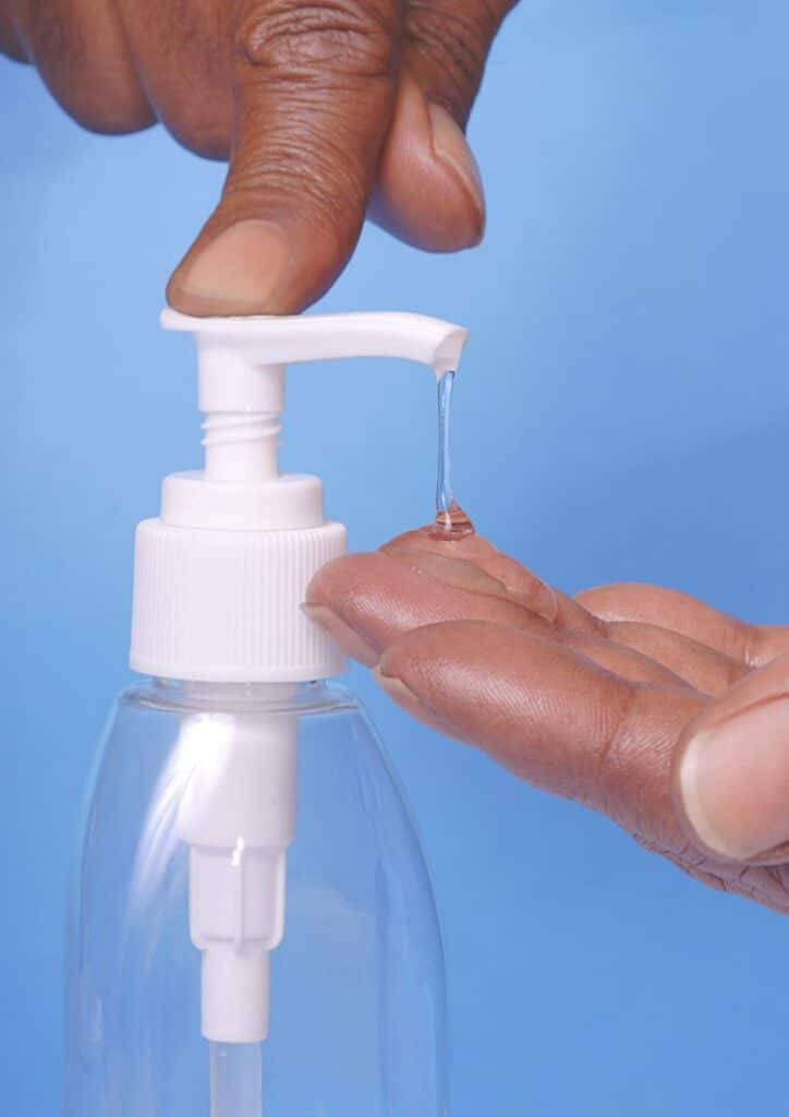 Hand pressing sanitizer pump, dispensing clear gel onto fingers against a blue background.