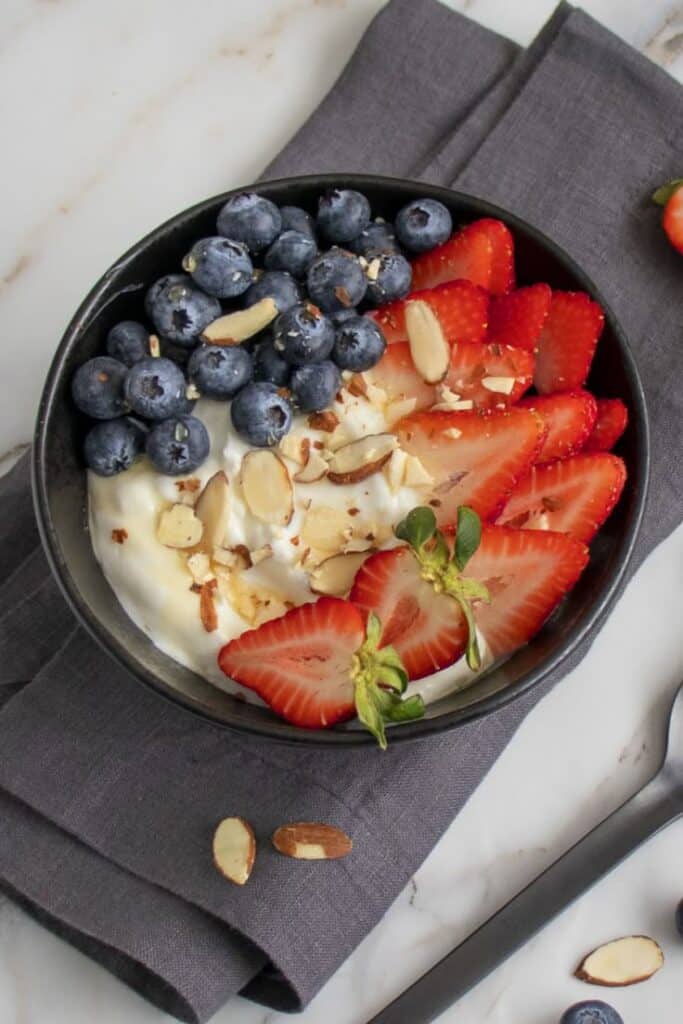 Yogurt bowl topped with fresh blueberries, strawberries, and sliced almonds on a dark napkin and marble surface.