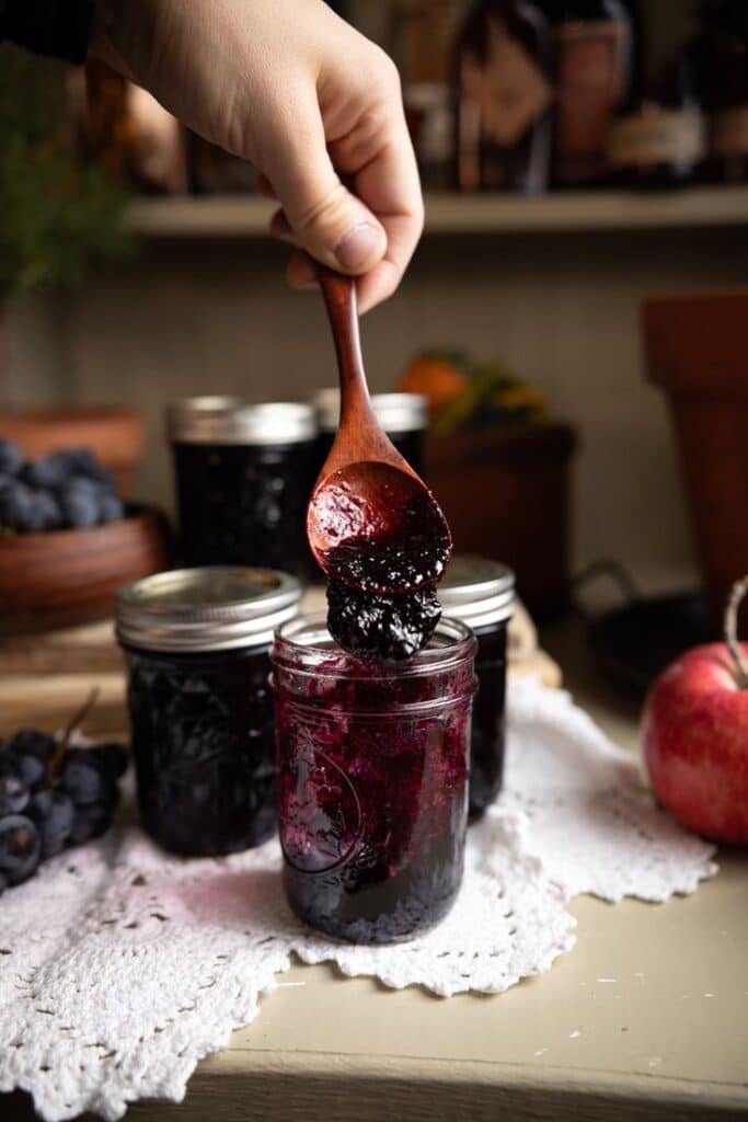 Hand scooping grape jelly from jar with wooden spoon, surrounded by jam jars and apple.