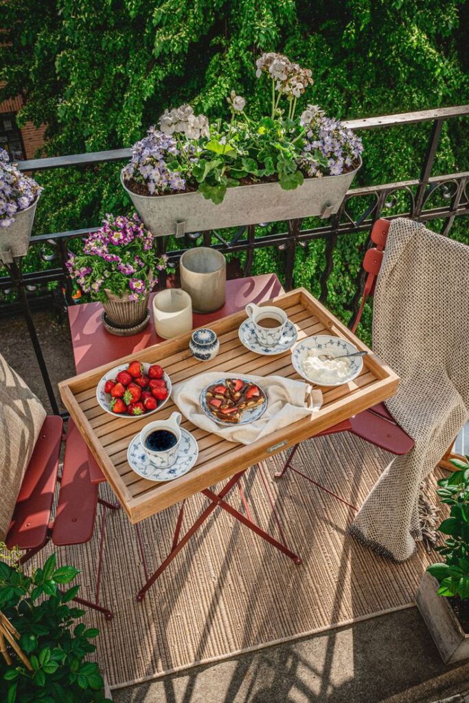 Cozy balcony breakfast with coffee, strawberries, and cake; surrounded by flowers and greenery.