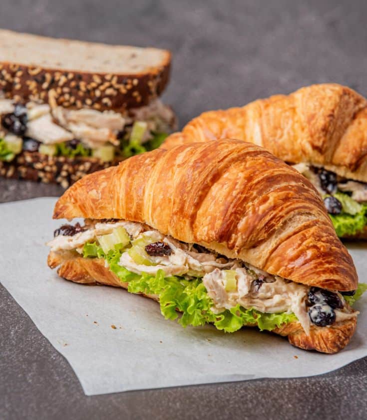 Chicken salad croissant sandwich with lettuce and cranberries on a parchment paper, with a sandwich in background.