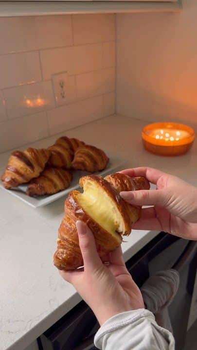 Cream-filled croissant being pulled apart in hands on kitchen counter with plate of croissants and candle in background.